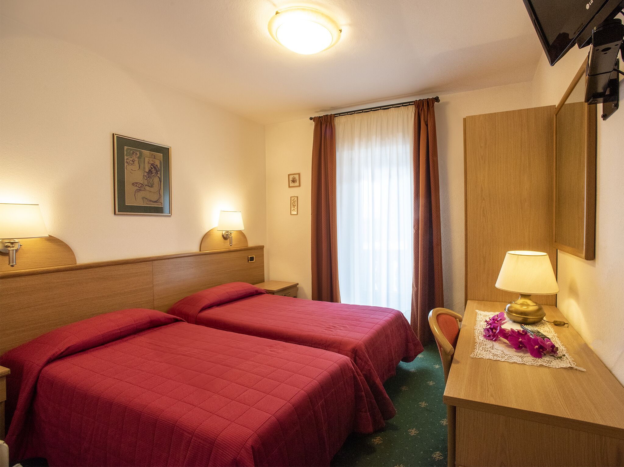 Ein Hotelzimmer mit zwei Einzelbetten, einem Schreibtisch mit Lampe, einem Spiegel und einem Fenster mit Vorhängen.