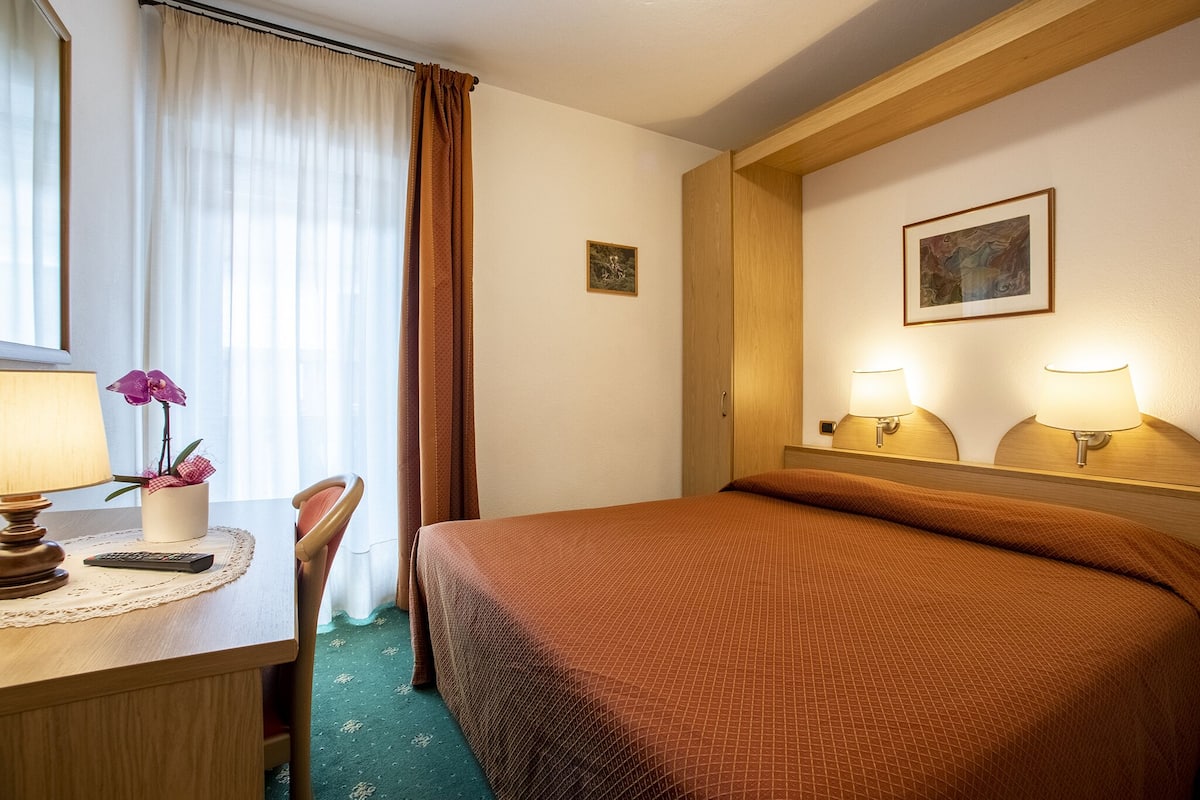 A hotel room with a bed, a desk with a lamp, a chair, and a window with curtains.
