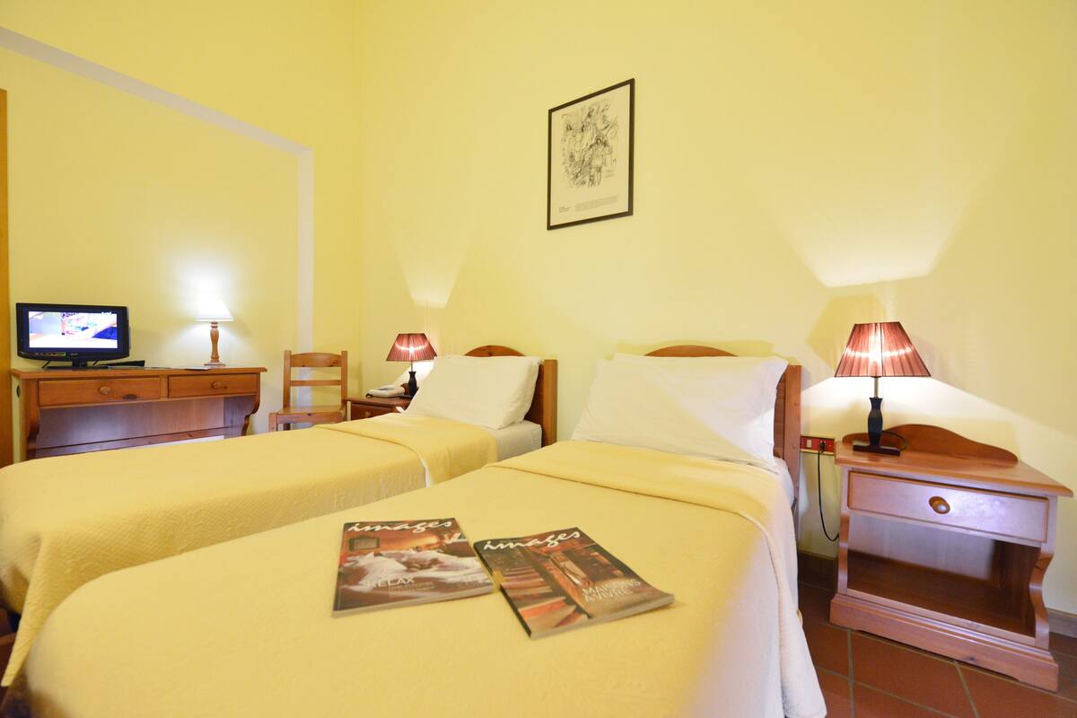 Quarto de hotel com duas camas, uma televisão sobre uma cômoda de madeira e duas luminárias de cabeceira.