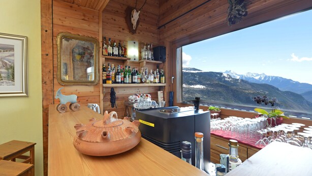 A wooden interior with a bar area, shelves with bottles, a large window with a mountain view, and a framed picture on the wall. - Hotel Maisonnette (Torgnon)