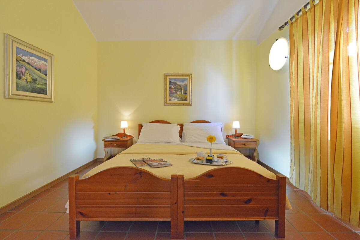 Quarto com cama de madeira, duas mesas de cabeceira com luminárias, um quadro de paisagem na parede e uma janela com cortinas.