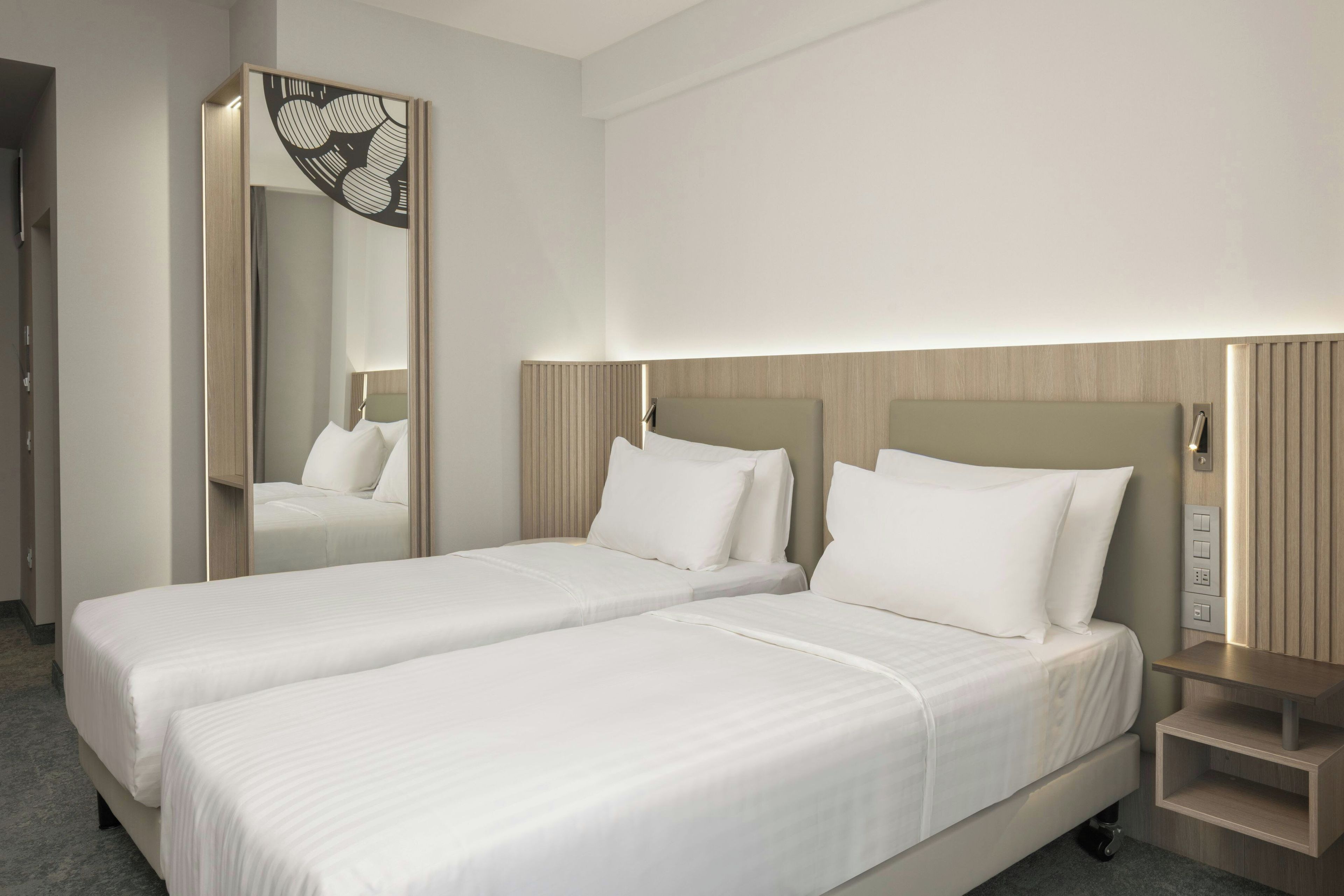A hotel room with two single beds, a mirror, a bedside table, and a wall-mounted light.