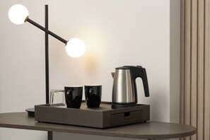 A modern table with a minimalist lamp, two black mugs, a clear glass, and a stainless steel kettle.