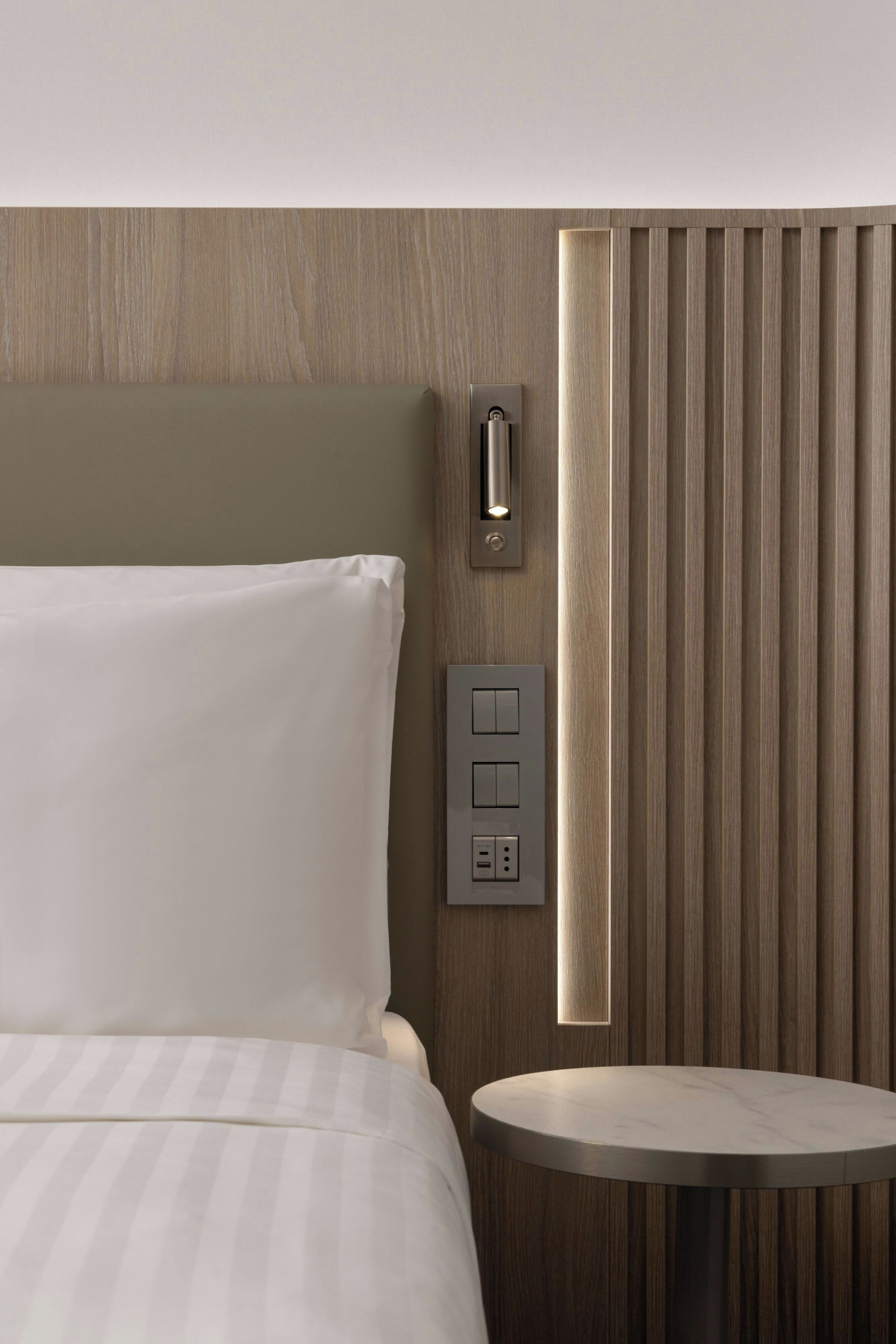 A hotel room with a bed, a bedside table, a wall-mounted control panel, and a vertical panel with a light.