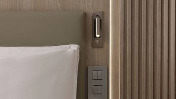 A hotel room with a bed, a bedside table, a wall-mounted control panel, and a vertical panel with a light.
