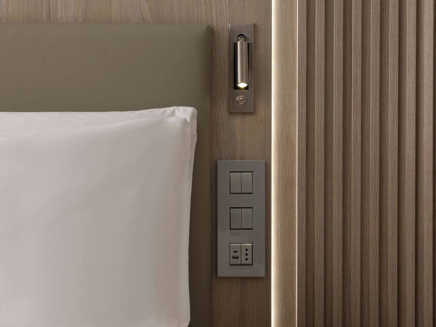 Habitación de hotel con cama, mesita de noche, panel de control en la pared y un panel vertical con luz.