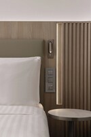 Una camera d'albergo con un letto, un comodino, un pannello di controllo a parete e un pannello verticale con una luce.