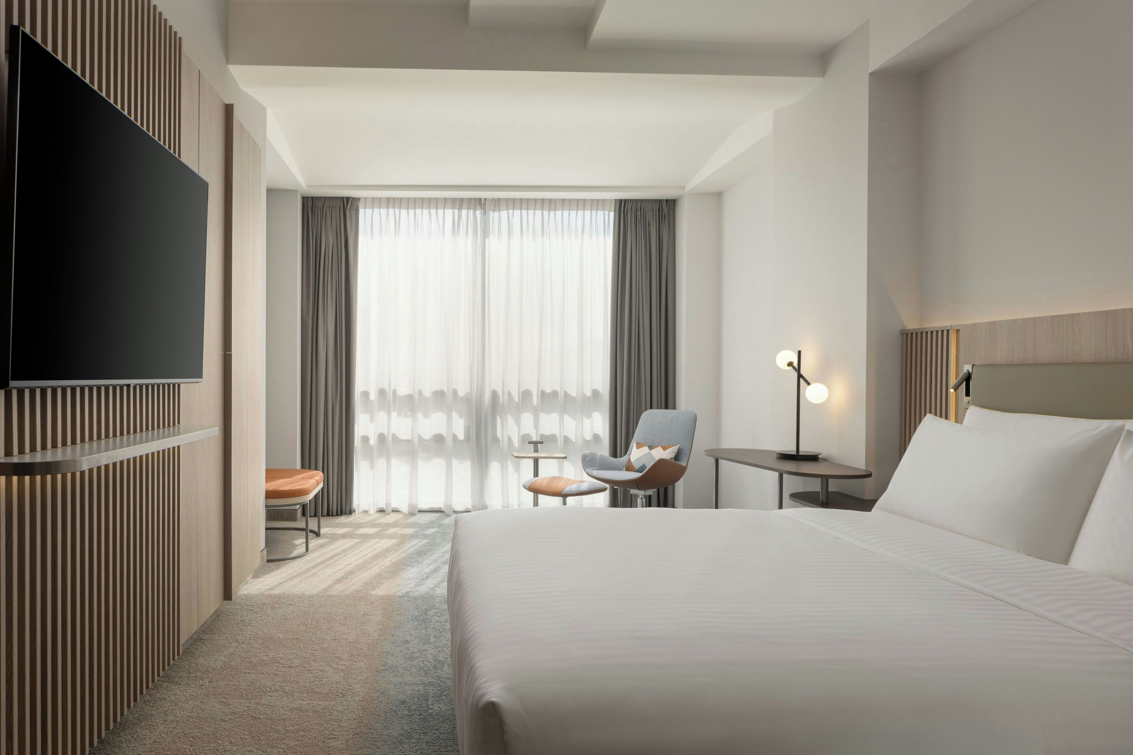 Habitación de hotel con una cama grande, un televisor de pantalla plana en la pared, un escritorio con una silla, una mesita con una lámpara y una ventana con cortinas.