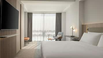 Habitación de hotel con una cama grande, un televisor de pantalla plana en la pared, un escritorio con una silla, una mesita con una lámpara y una ventana con cortinas.