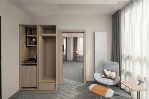 Habitación de hotel con armario, zona de estar con sillón y reposapiés, una mesita pequeña y un ventanal con cortinas.