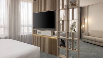 A modern hotel room with a large flat-screen TV mounted on a wooden panel, a bed with white bedding, a sofa, and a carpeted floor.