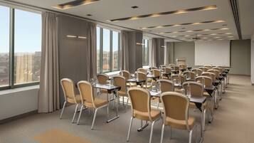 A conference room with rows of tables and chairs, large windows, and a projector screen.