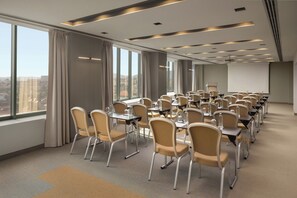 A conference room with rows of tables and chairs, large windows, and a projector screen.