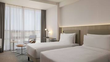 A hotel room with two beds, a desk, a chair, and a lamp.