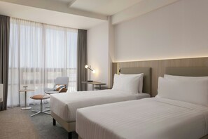 A hotel room with two beds, a desk, a chair, and a lamp.