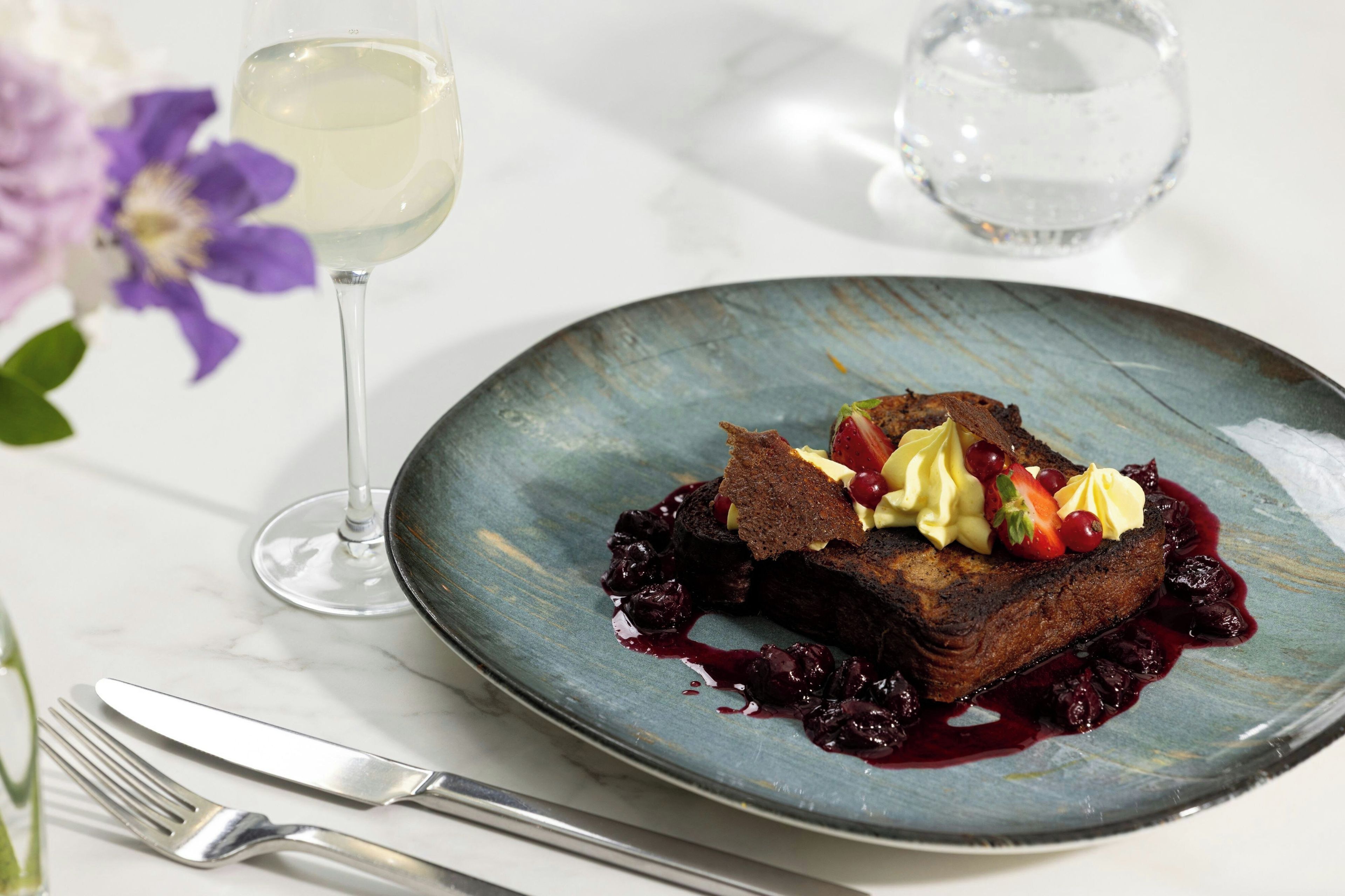 A dessert plate with a dark cake, fruit, and cream on a blue plate, accompanied by a glass of white wine and a fork on a marble surface.
