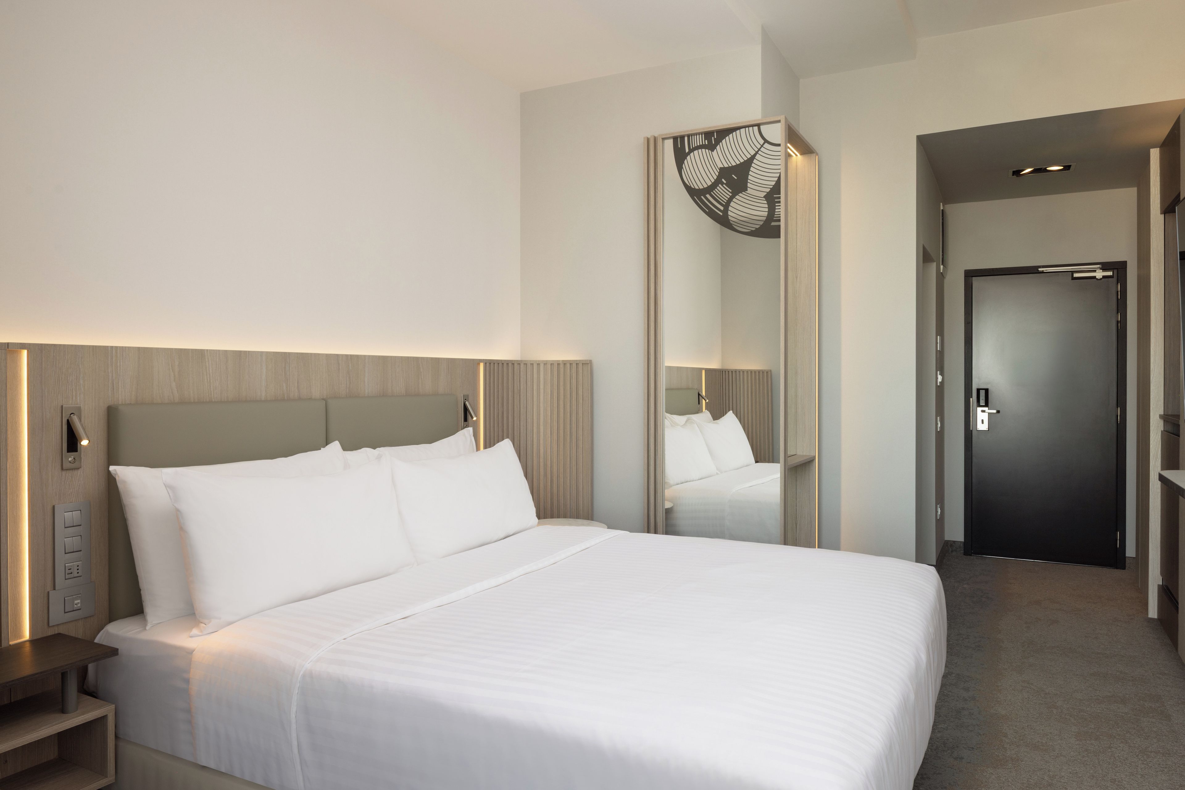 A modern hotel room with a large bed, bedside controls, a mirror, and a door leading to another room.