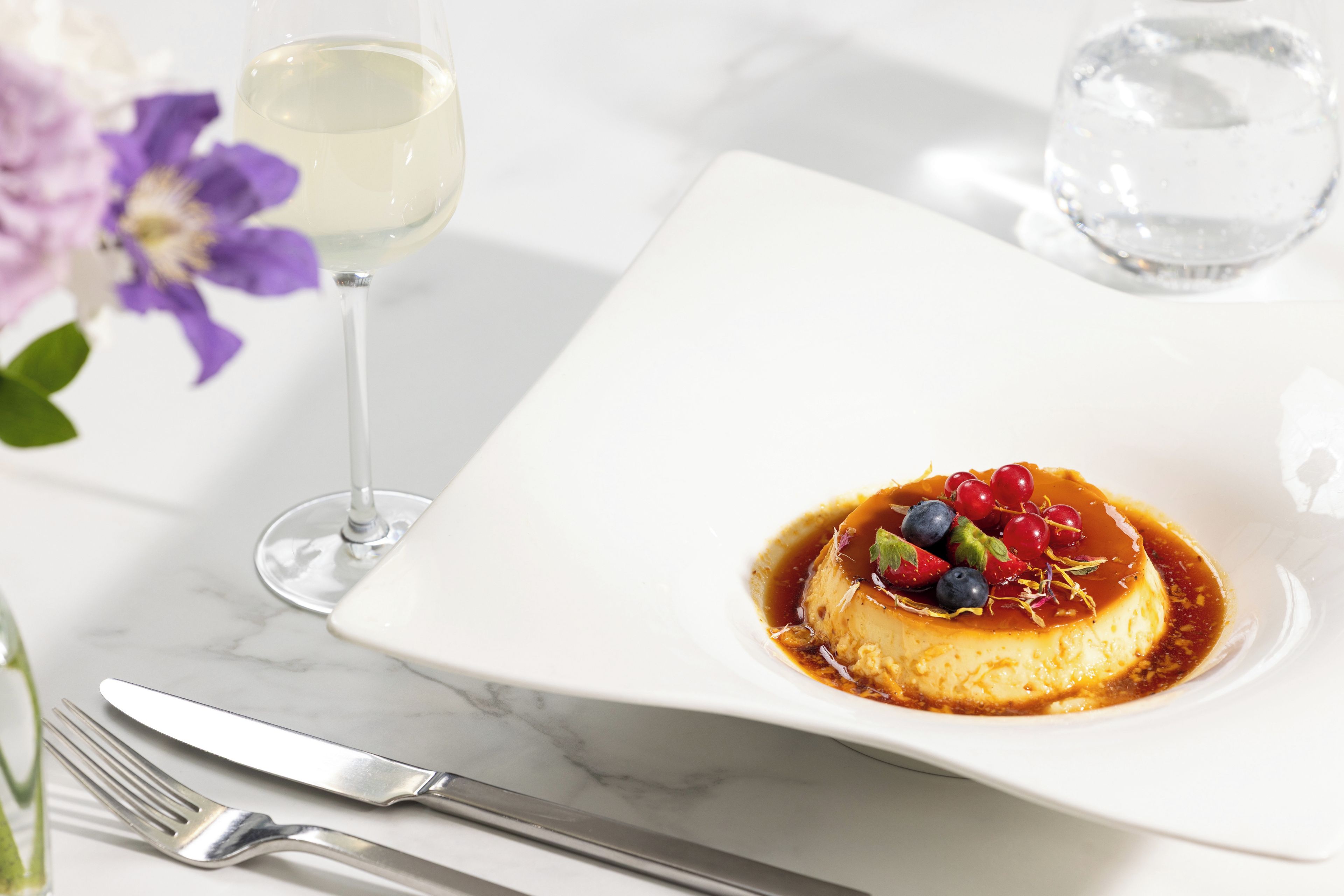 Un dessert servi sur une assiette carrée, un verre de vin blanc et une fourchette posée sur une table en marbre.