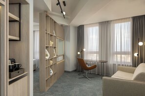 A modern hotel room with a wooden bookshelf, a round table with a chair, and large windows with curtains.