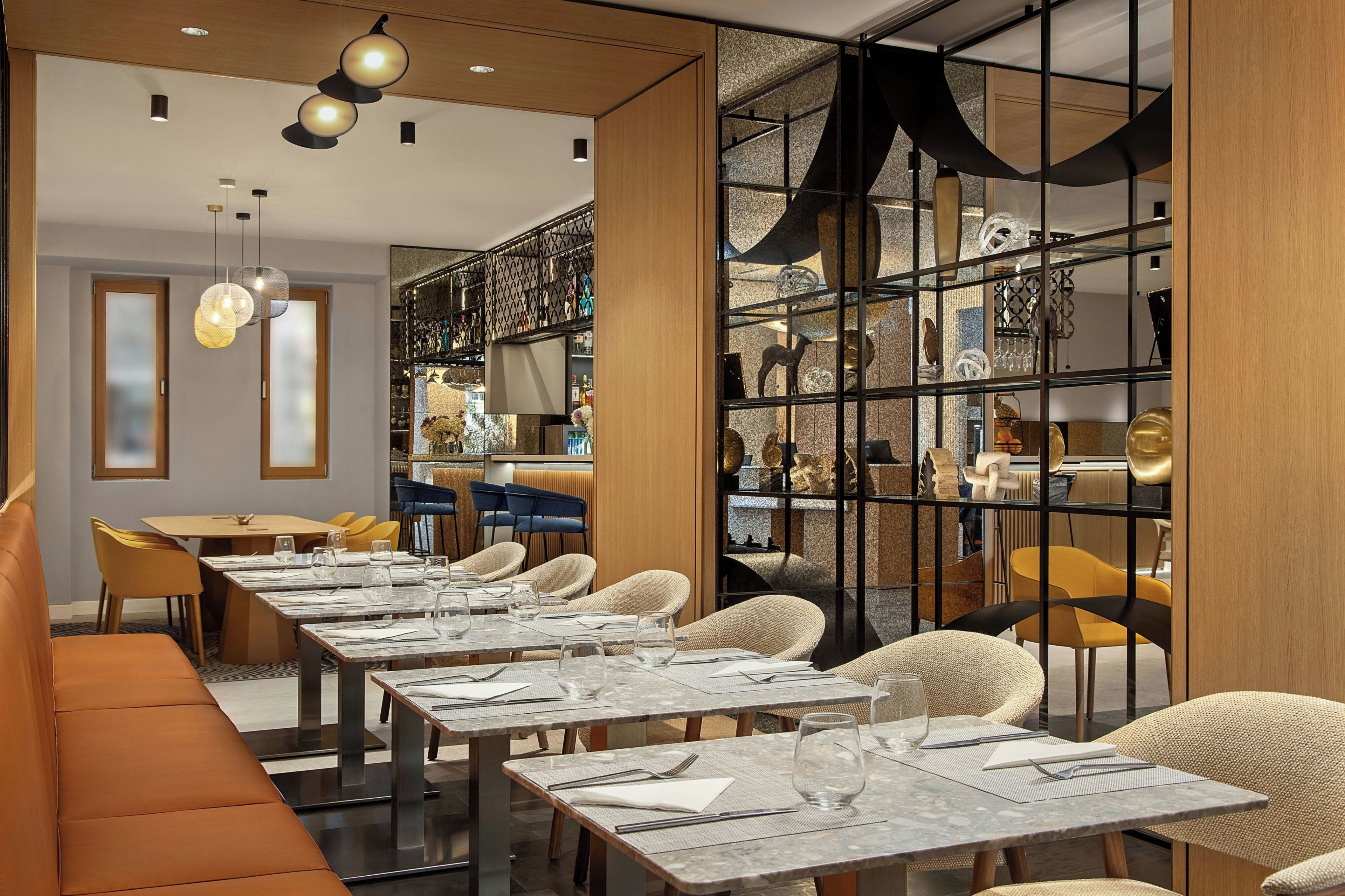A modern dining area with multiple tables and chairs, a bar area with shelves of bottles, and a decorative wall with a geometric pattern.