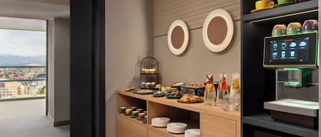 A modern hotel room with a breakfast bar, a coffee machine, and a view of the city.