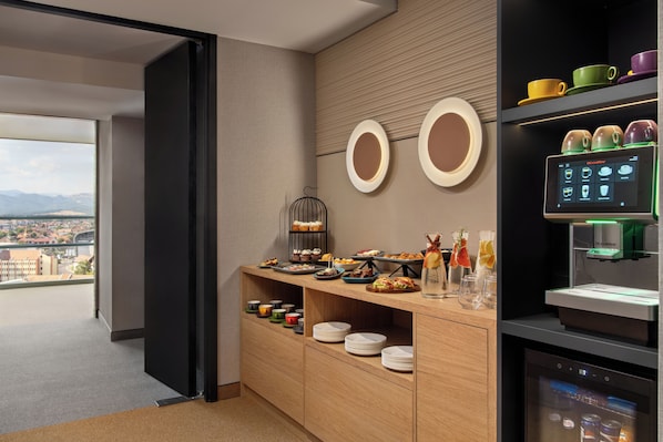 A modern hotel room with a breakfast bar, a coffee machine, and a view of the city.