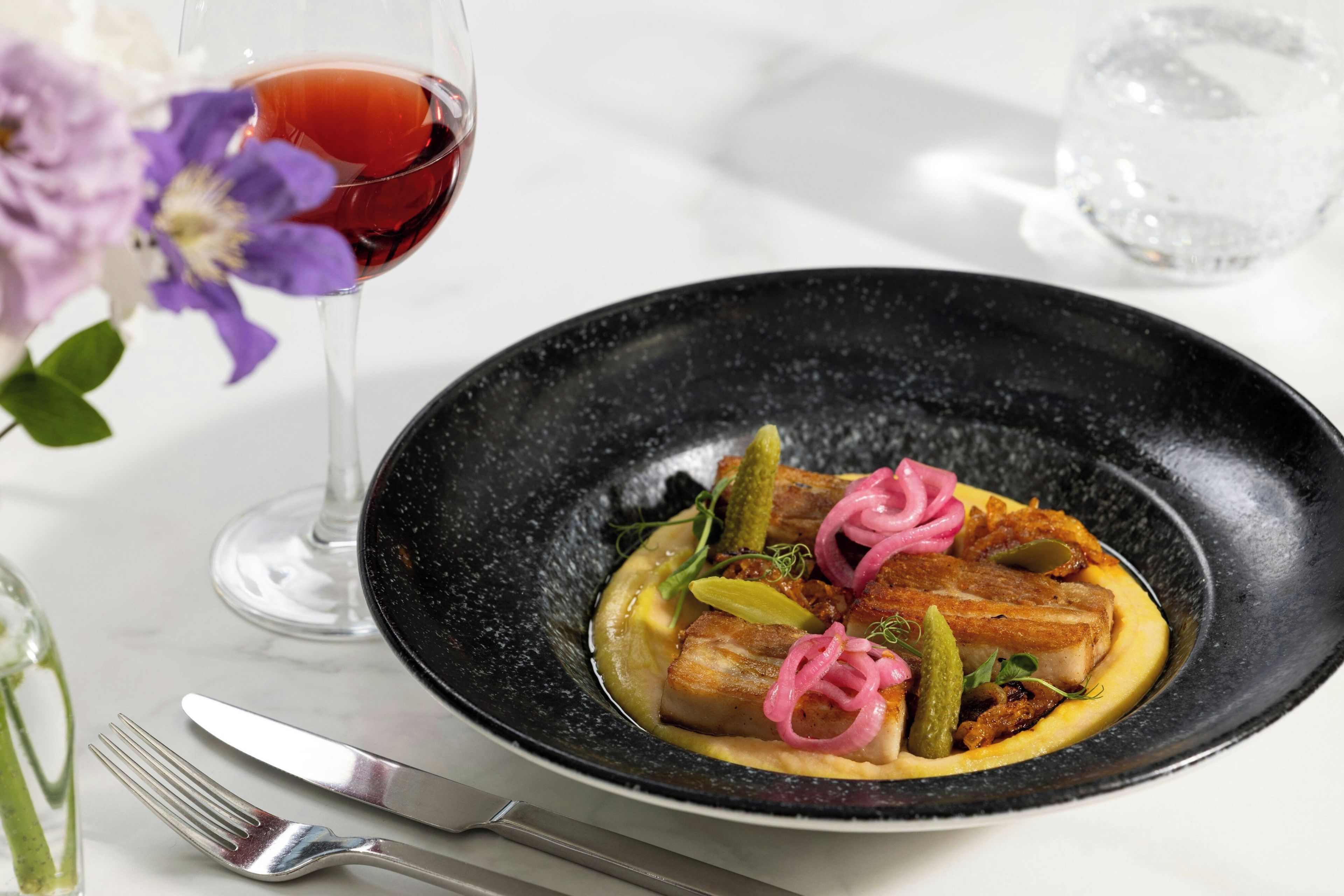A black plate with food, a glass of red wine, and a fork on a table.