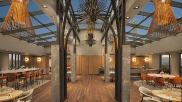 A modern dining area with high ceilings, exposed beams, and a central chandelier. Tables are set for dining with chairs.