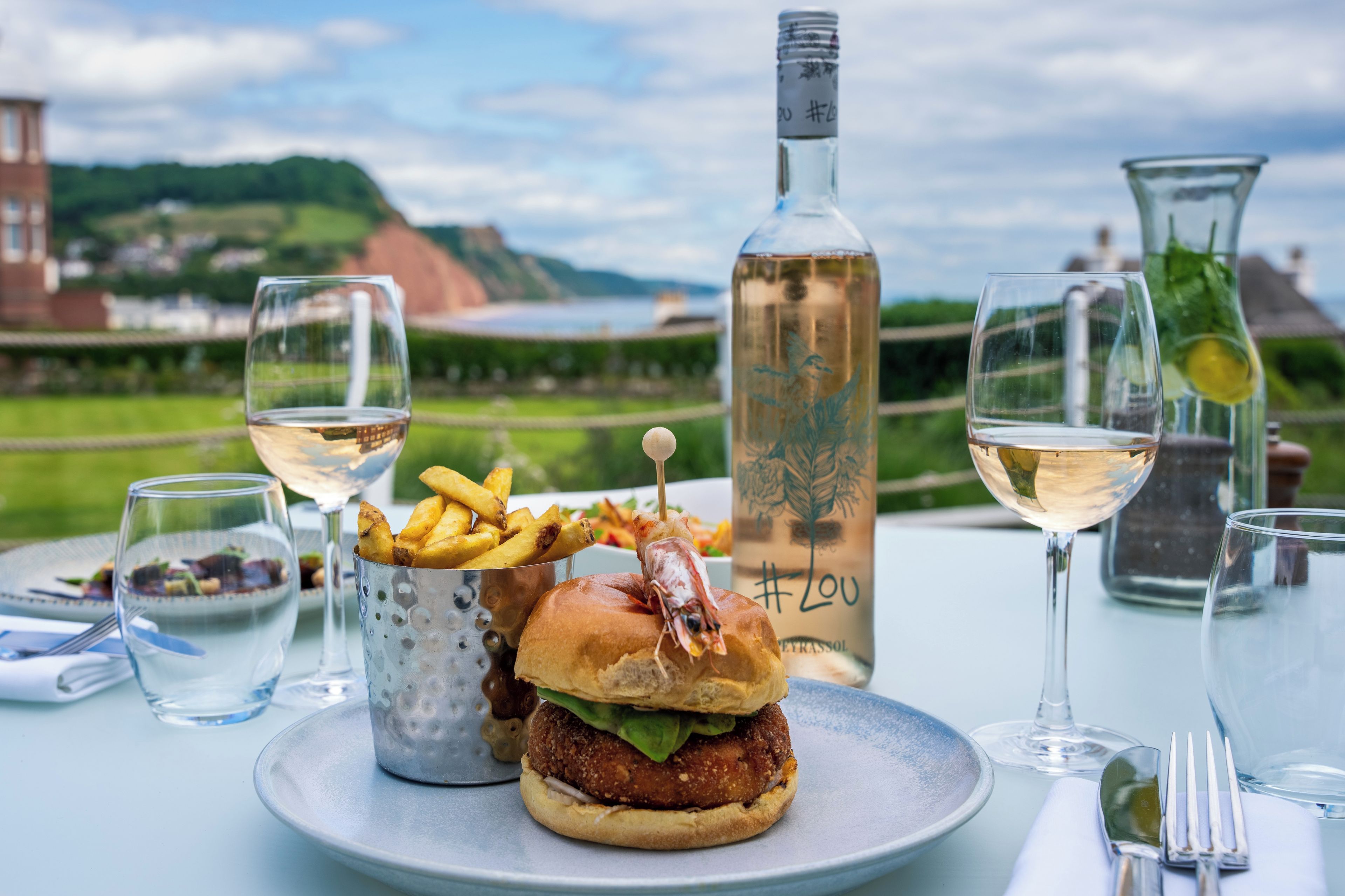 A dining table with a burger, fries, and a bottle of wine, set against a scenic outdoor background.