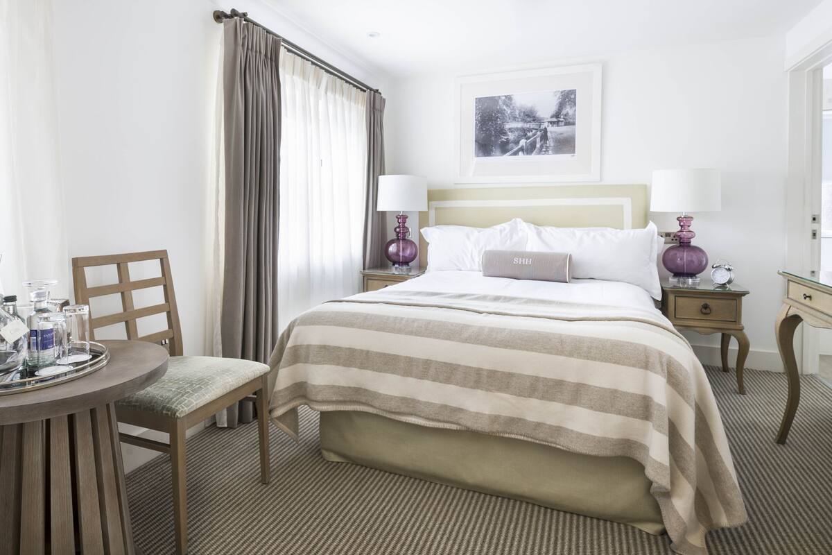 Une chambre d’hôtel comprenant un lit, deux tables de chevet, une chaise, une petite table avec un plateau, et une fenêtre avec des rideaux.