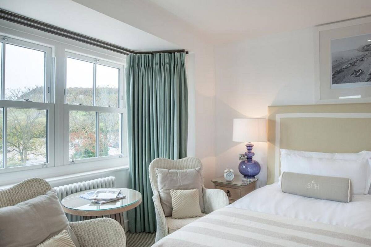 Une chambre d’hôtel avec un lit, une chaise, une table, une lampe et une fenêtre avec des rideaux.