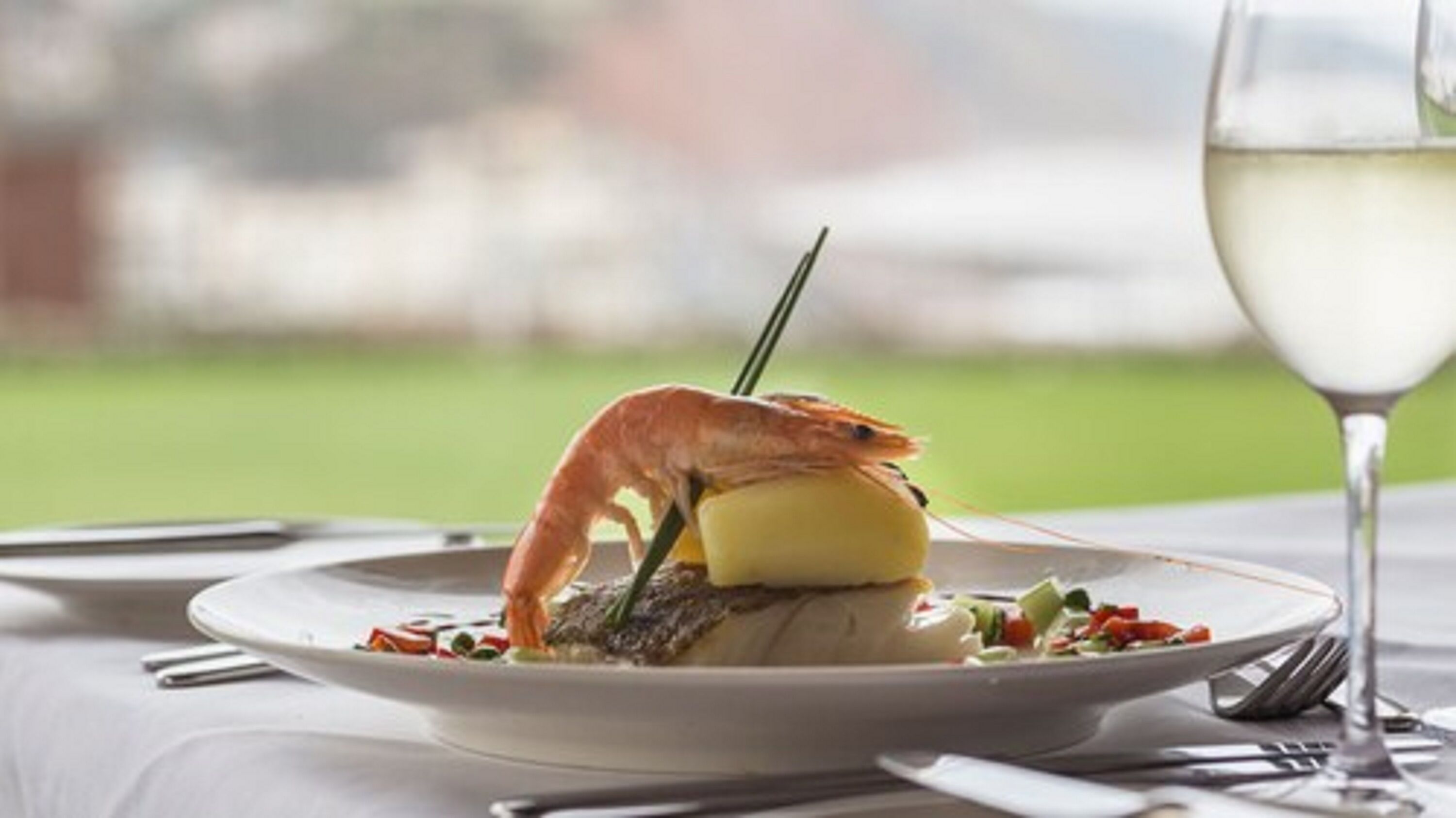 A plate with a shrimp, a yellow item, and a green garnish on a table with a glass of white wine.