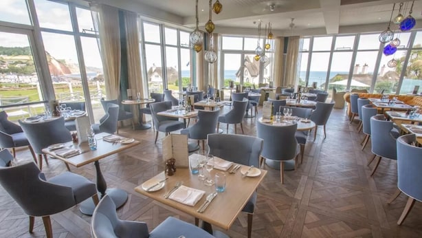 A restaurant with tables set for dining, large windows offering a view of the sea and hills, and hanging light fixtures.
