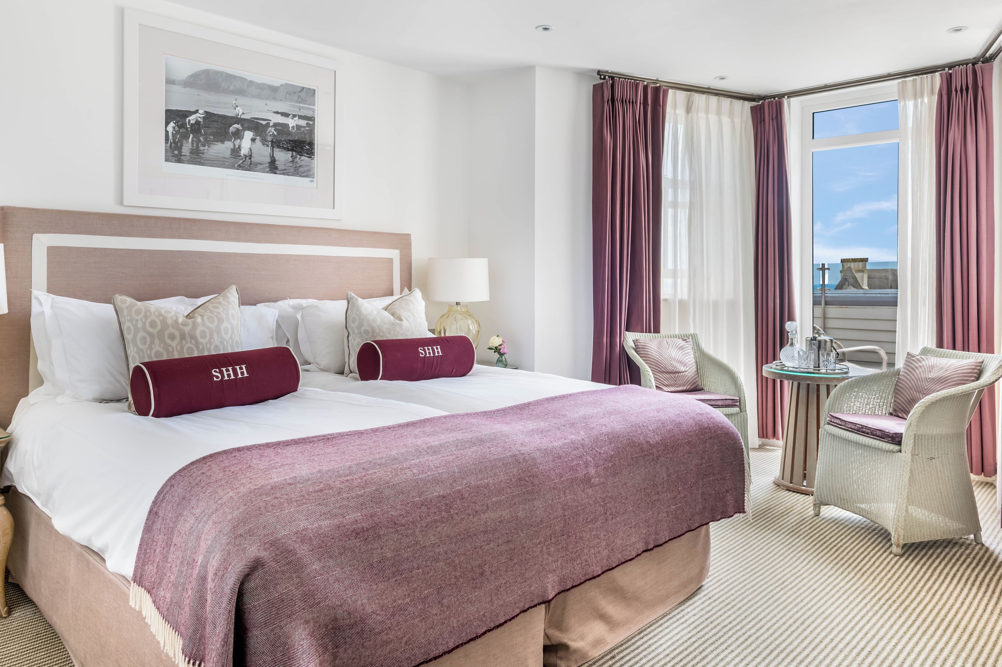 A hotel room with a large bed, two pillows with 'SHH' on them, a side table with a lamp, a chair, and a window with curtains.