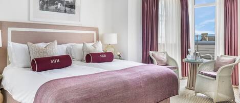 A hotel room with a large bed, two pillows with 'SHH' on them, a side table with a lamp, a chair, and a window with curtains.