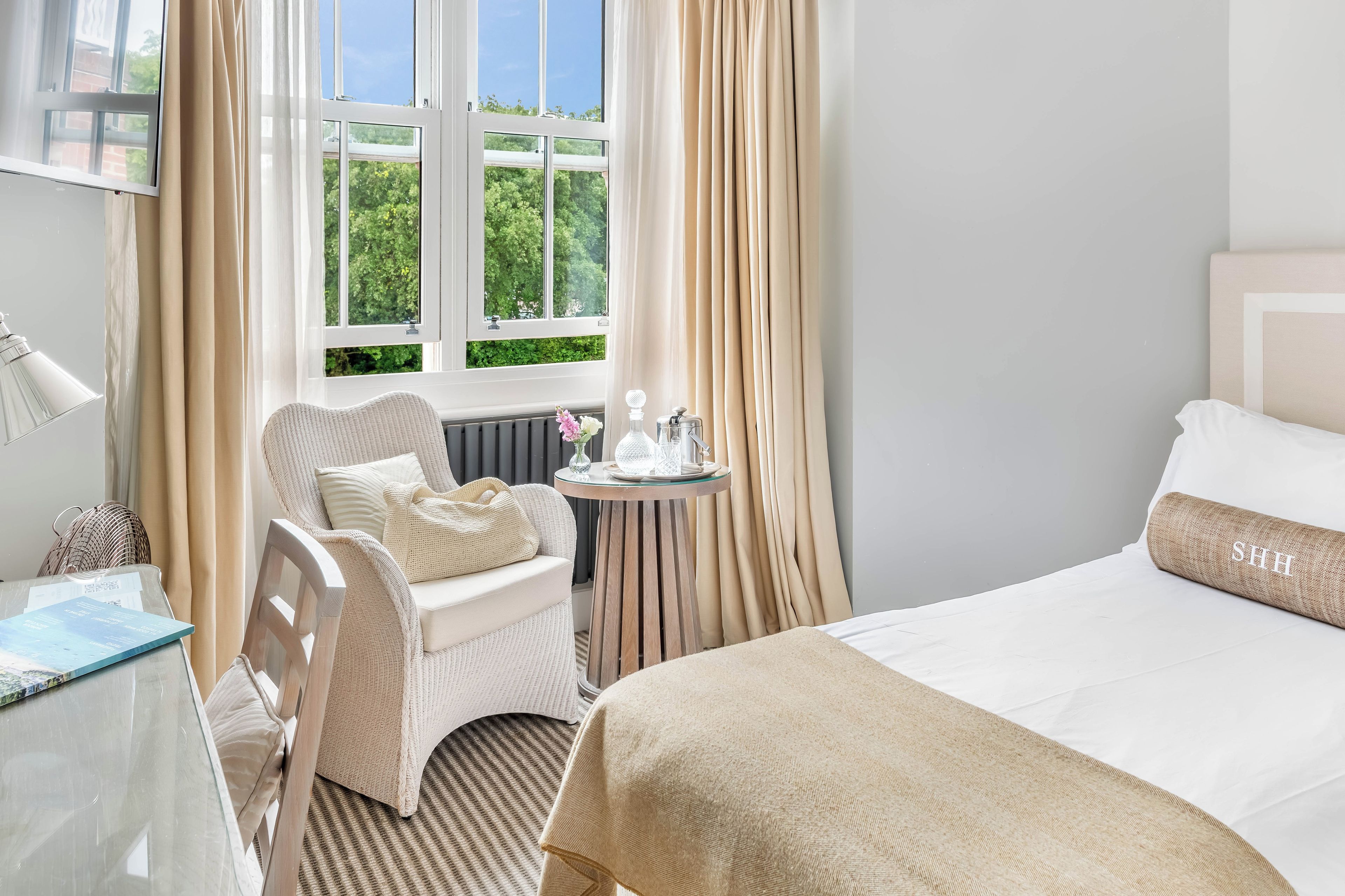 Habitación de hotel con una cama, una silla, una mesita con un florero y una ventana con cortinas.