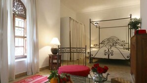 Un dormitorio con cama con dosel, una mesita con fruta y un florero, una lámpara y una ventana con cortinas.