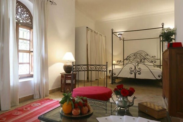 Un dormitorio con cama con dosel, una mesita con fruta y un florero, una lámpara y una ventana con cortinas.