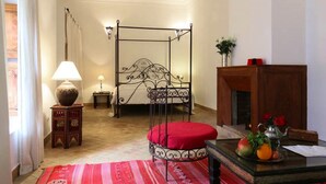 Un dormitorio con una cama con dosel, una mesita de noche de madera con una lámpara, un reposapiés rojo, una pequeña mesita con un jarrón y un frutero sobre una mesa de madera.