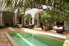 Un área de piscina con suelo de baldosas, rodeada de plantas en macetas y mobiliario.