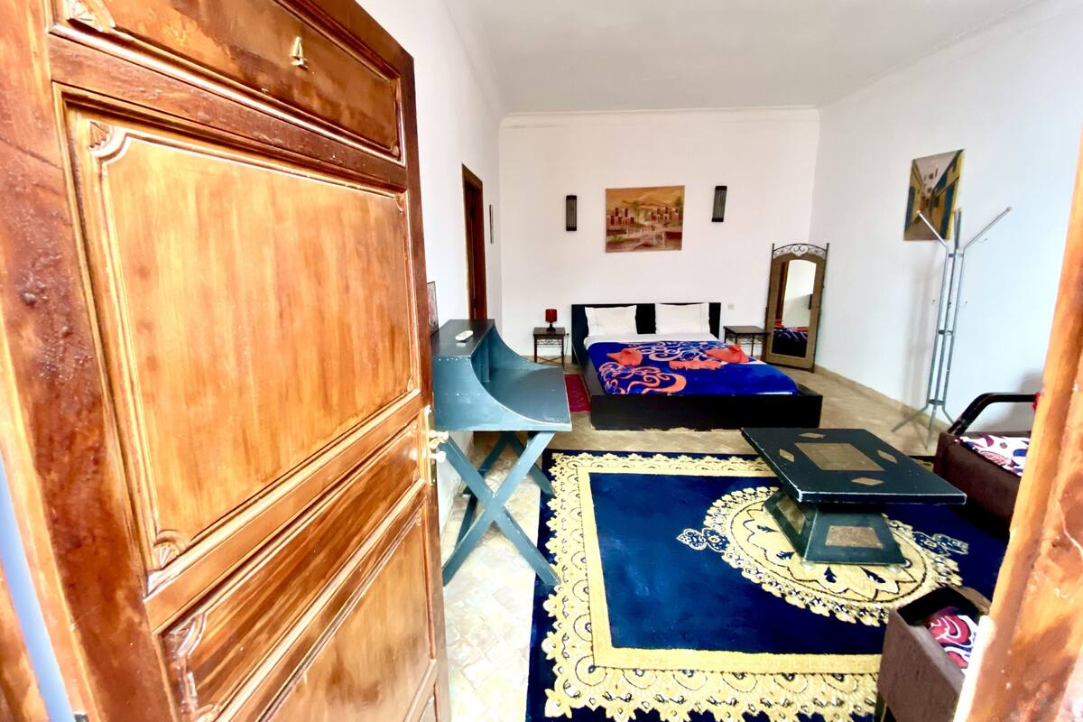 A bedroom with a wooden wardrobe, a bed with a blue and orange bedspread, a blue and gold patterned rug, and a small table with a chair.