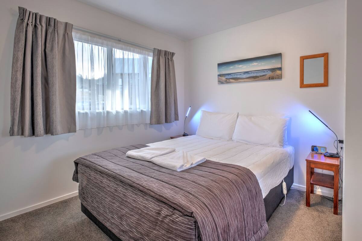 A bedroom with a bed, a bedside table, a nightstand, a lamp, a painting, and a window with curtains.