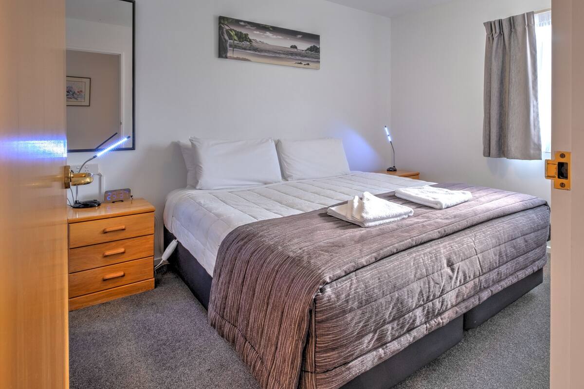 A bedroom with a bed, a bedside table with a lamp, a nightstand, a wall-mounted painting, and a window with curtains.