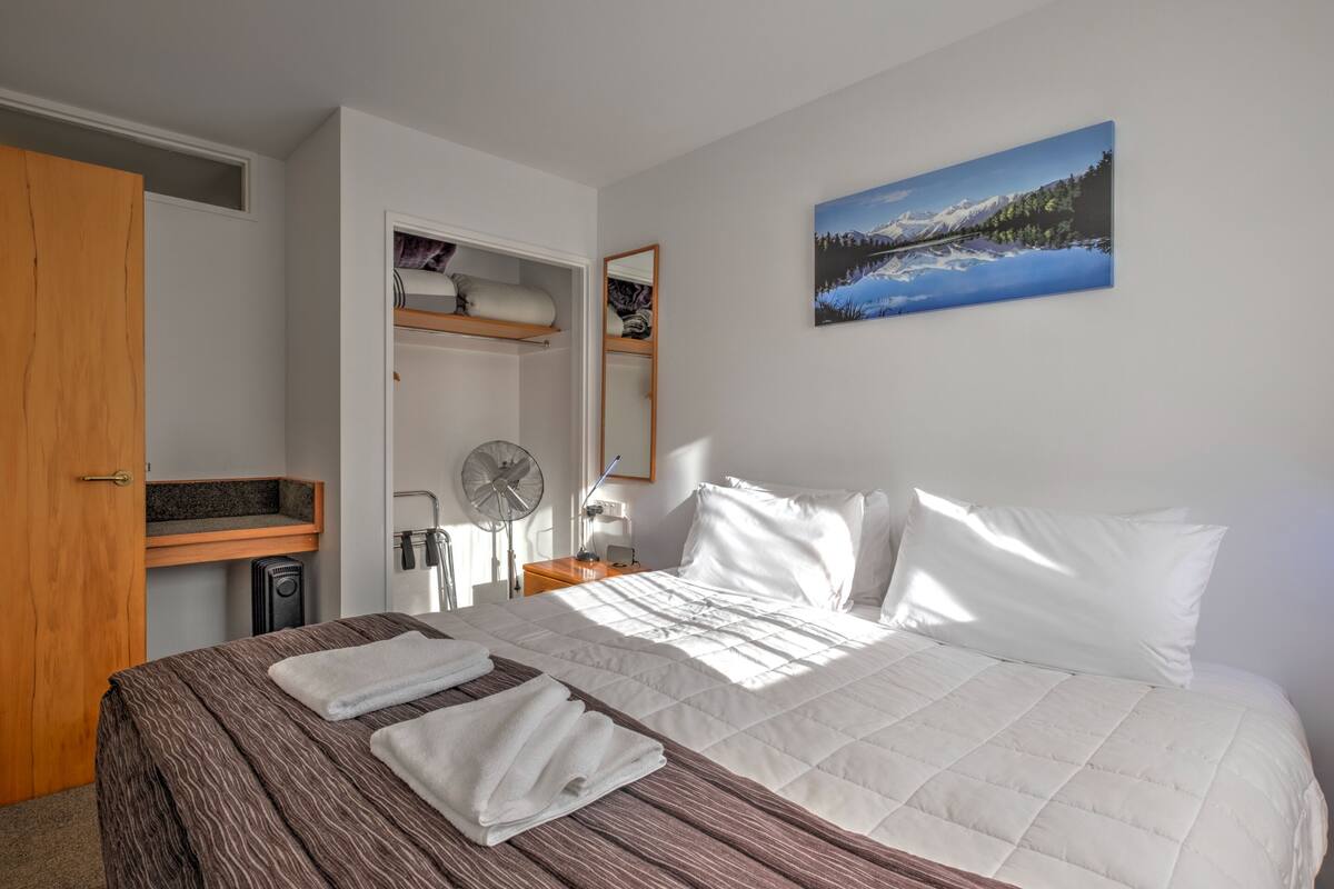 A neatly made bed with white linens and a brown striped blanket, a wooden nightstand with a lamp, a wall-mounted painting of a mountain landscape, and a small shelf with personal items.
