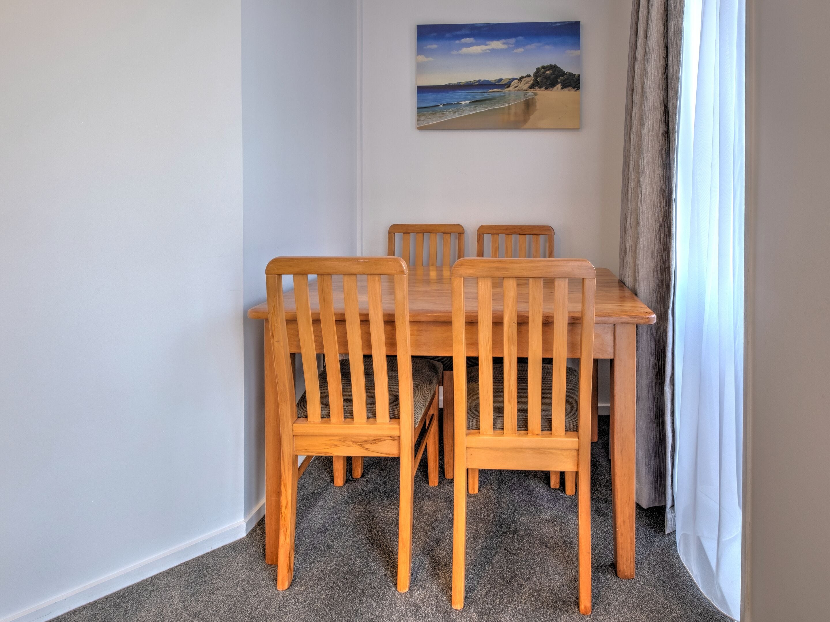 A dining area with a wooden table and chairs, carpeted floor, and a beach scene painting on the wall.