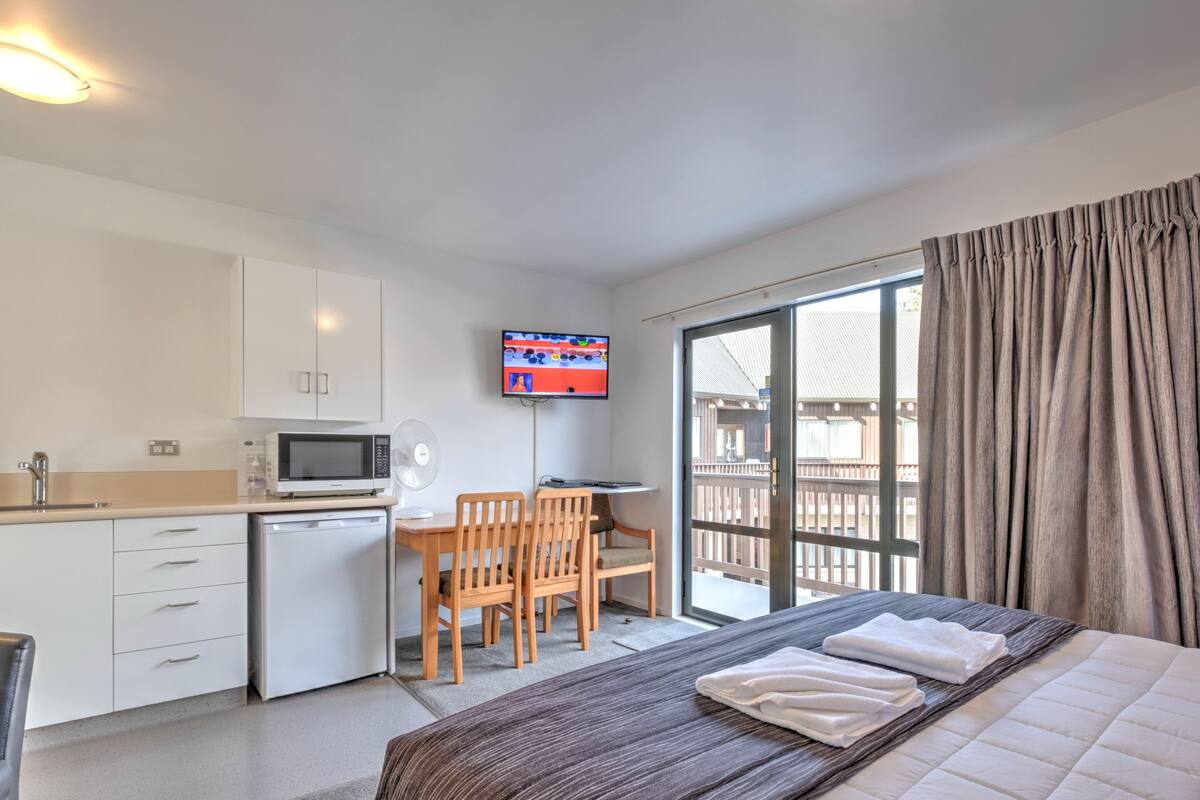 A hotel room with a bed, a desk with a chair, a TV, and a balcony with curtains.