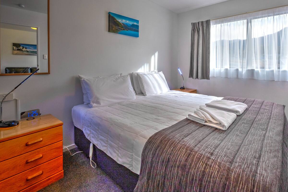 A neatly arranged bedroom with a bed, bedside table, dresser, and a painting on the wall.