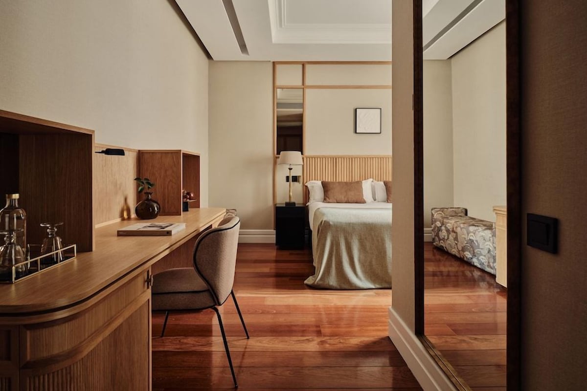 Une chambre d’hôtel avec un bureau en bois, une chaise, un lit recouvert d’une courtepointe blanche et une table de chevet avec une lampe.