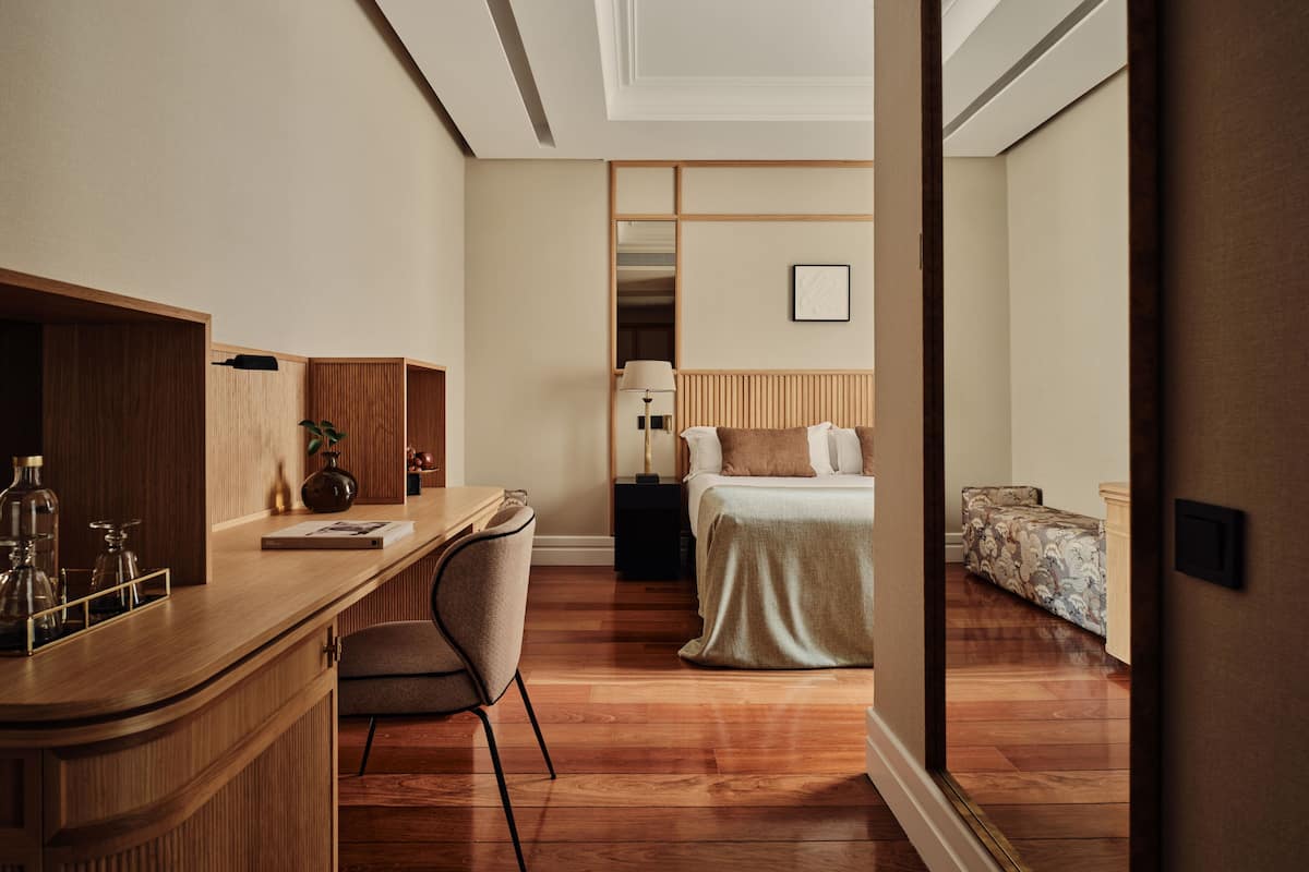 Une chambre d’hôtel avec un bureau en bois, une chaise, un lit avec une tête de lit, une table de chevet et un petit canapé.