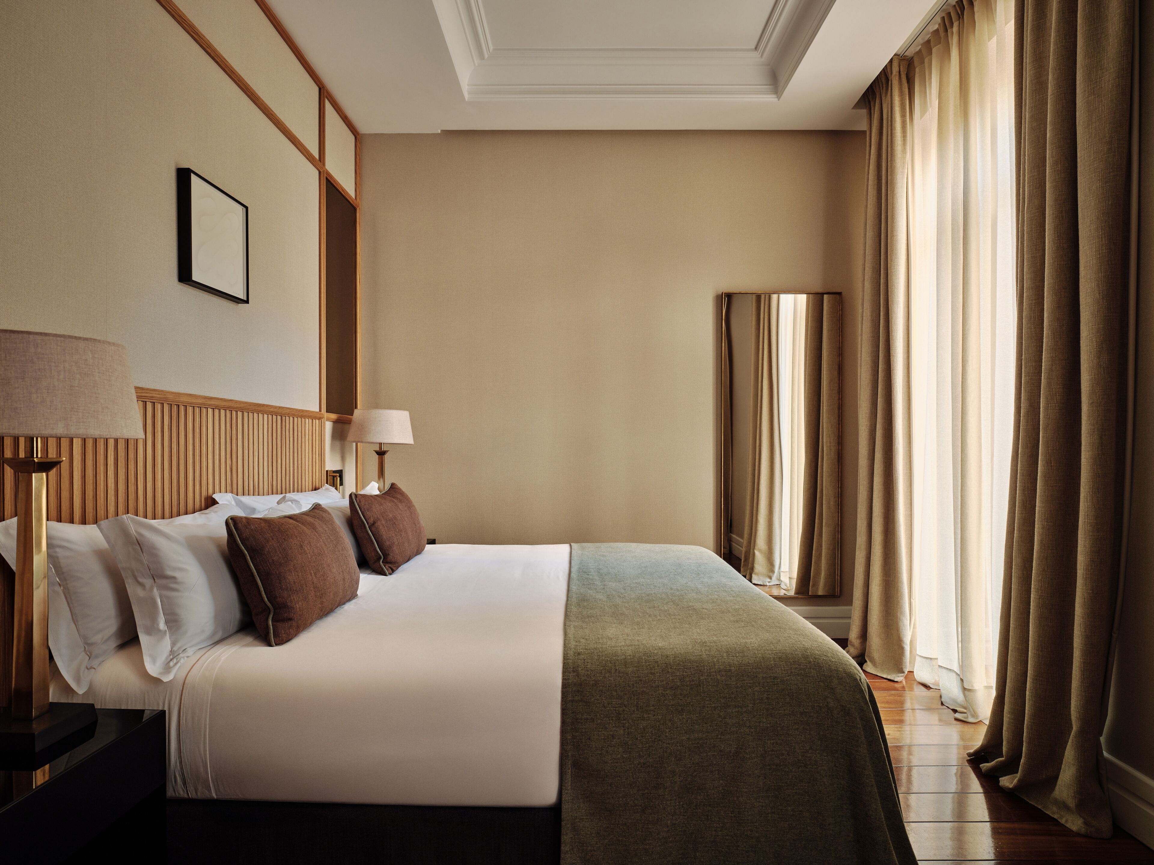 Habitació d'hotel amb un llit ben fet, una taula de nit amb una làmpada, una finestra gran amb cortines i un panell de capçal de fusta.